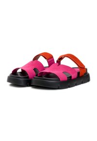 Paire de sandales en daim rose avec semelles épaisses noires et brides de cheville réglables orange, orientées vers l'avant sur un fond blanc.