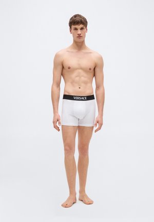 Noore mees seisab paljajalu, kandes valgeid Versace bokseraluspükse musta vöökohaga, ühetaolise heleda tausta ees.