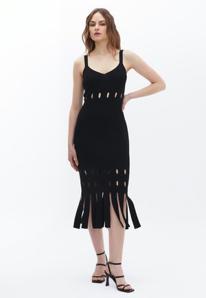 Robe noire tricotée avec une silhouette ajustée, présentant des découpes circulaires le long du corsage et un ourlet à franges, complétée par des talons noirs à lanières.