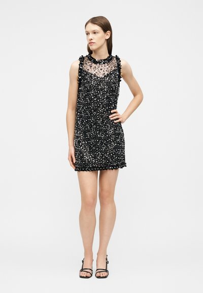 Femme portant une mini robe noire sans manches avec des ornements floraux et un décolleté transparent, assortie à des sandales à talons hauts noires à lanières.