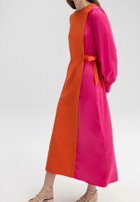 Femme portant une longue robe avec un panneau avant orange et des manches et côtés rose vif, avec une ceinture à la taille et des talons beige à brides.