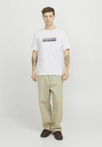 Witte katoenen T-shirt met "JACK & JONES" in blauw en rood bedrukt. Gecombineerd met lichtbeige jeans en bruine leren schoenen.