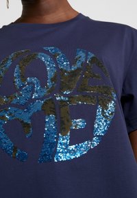 Camiseta azul marino con un gran gráfico de lentejuelas "LOVE ME" en tonos de azul y verde, con una textura de tela suave.