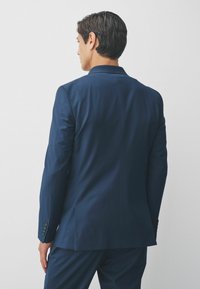 Marineblauwe colbert met een getailleerde pasvorm, pieklapel, gladde textuur en vierknopenmanchetten, weergegeven vanaf de achterkant.