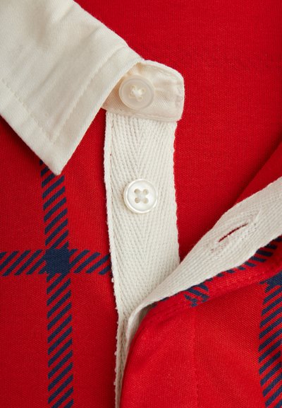 Κόκκινη πόλο μπλούζα με λευκό γιακά, διακοσμημένη με ναυτικές καρό λεπτομέρειες. Κλείσιμο με κουμπιά και υφασμάτινη λωρίδα δίπλα στα κουμπιά.