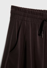 Tmavě hnědé měkké kalhoty z tkaniny s elastickým pasem a šňůrkou, které mají boční kapsy a hladký povrch.
