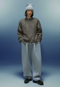 Šedý pletený sveter s textúrovaným povrchom, ktorý je navrstvený na kockovanej košeli. Voľné šedé teplákové nohavice a čierne topánky dotvárajú celý outfit.