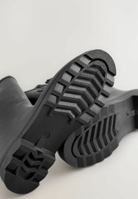 Bottes en caoutchouc noir avec une semelle texturée dotée de profonds crampons et d'une surface supérieure lisse. Les semelles présentent un motif à crampons pour une meilleure adhérence.