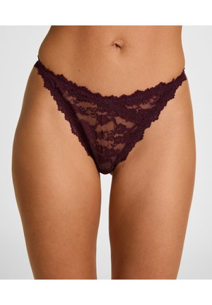 Frau trägt dunkelvioletten Spitzenbikini mit Blumenmuster, zeigt Taille und obere Oberschenkel vor einfarbigem Hintergrund.