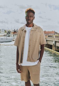 Jeune homme aux cheveux bouclés portant une chemise à manches courtes texturée beige et un short par-dessus un t-shirt blanc, debout près d'une marina avec des bateaux et de l'eau.