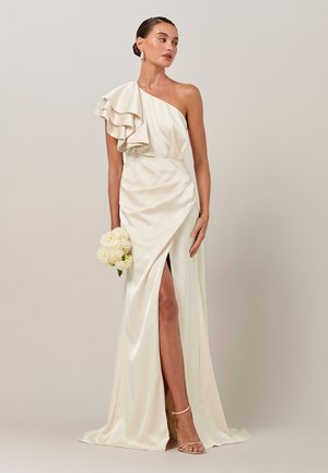 Žena v krémových saténových šatách s jedným ramenom, riaseným rukávom a rozparkom na stehne, drží biely kyticu kvetov, stojaca pred neutrálnym pozadím.