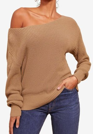 Femme portant un pull en maille ample de couleur camel, une épaule découverte, associée à un jean bleu foncé, main dans la poche.