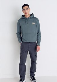 Zielona bawełniana bluza z kapturem z beżowym logo, z przednią kieszenią; dopasowana do szarych spodni cargo z wieloma kieszeniami; czarno-białe sneakersy.