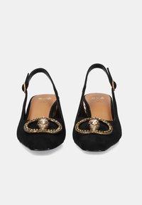 Tacones slingback de ante negro con punta redondeada, adornados con un detalle ornamental en tono dorado y pequeños acentos circulares.