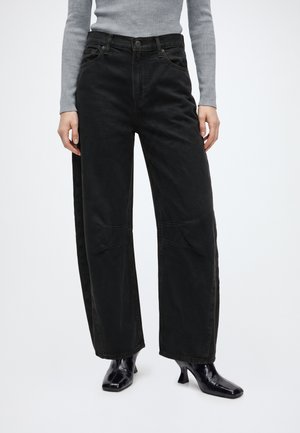 Person iført højtaljede løse sorte jeans, grå ribbet langærmet top og skinnende sorte ankelstøvler med hæl på hvid baggrund.