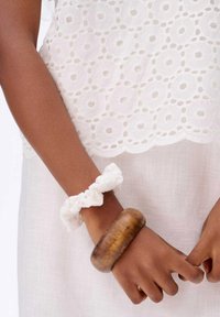 Biała koronkowa bluzka z okrągłymi wycięciami, biały materiałowy gumka do włosów i brązowy drewniany bransoletka na brązowej skórze nadgarstka. Jasne bawełniane tło.