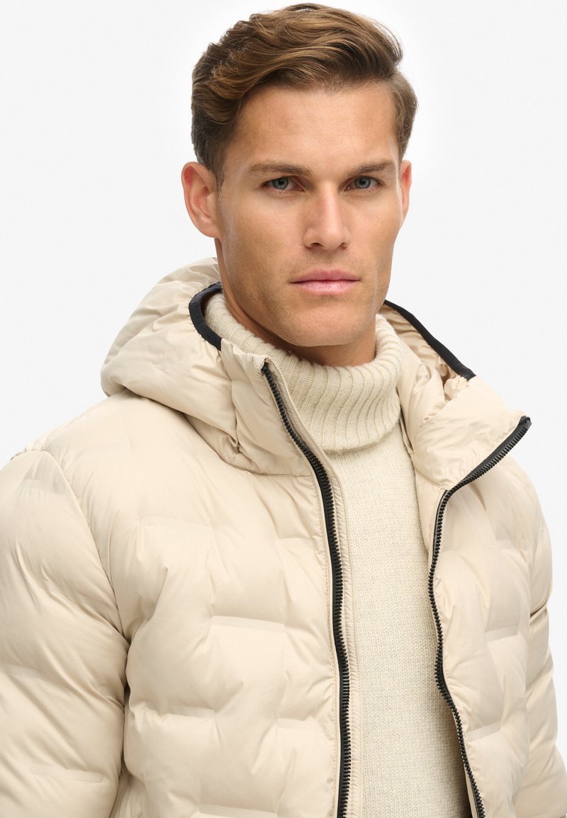 Beige donsjas met een ritssluiting aan de voorkant, een capuchon en een geribbelde gebreide trui eronder. Gelaagd stof met een zichtbaar doorgestikt patroon en donkere accenten.