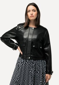 Schwarze, cropped Lederjacke mit zwei vorderen Taschen, Druckknöpfen und einer glatten Oberfläche. Kombiniert mit einem schwarz-weißen Rock mit Zickzackmuster.