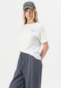 Hvit bomulls T-skjorte med grønn logo "THE MOON" på brystet. Bært med mørk blå teksturerte, løstsittende bukser og en slitt grå caps.