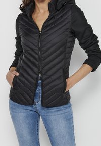 Femme portant une veste matelassée noire avec capuche par-dessus un haut noir à manches longues et un jean bleu, mains dans les poches de la veste, fermeture éclair avant fermée.