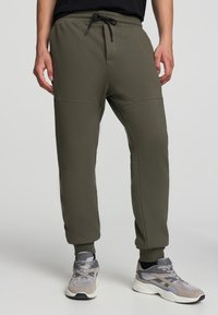 Oliwkowe dresy z elastycznym pasem i regulowanym sznurkiem. Posiadają zwężające się nogawki oraz boczne szwy z teksturowanymi panelami.