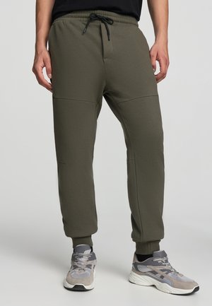 Pantaloni jogger verde oliva con vita elastica e coulisse regolabile. Presentano gambe affusolate e cuciture laterali con pannelli testurizzati.