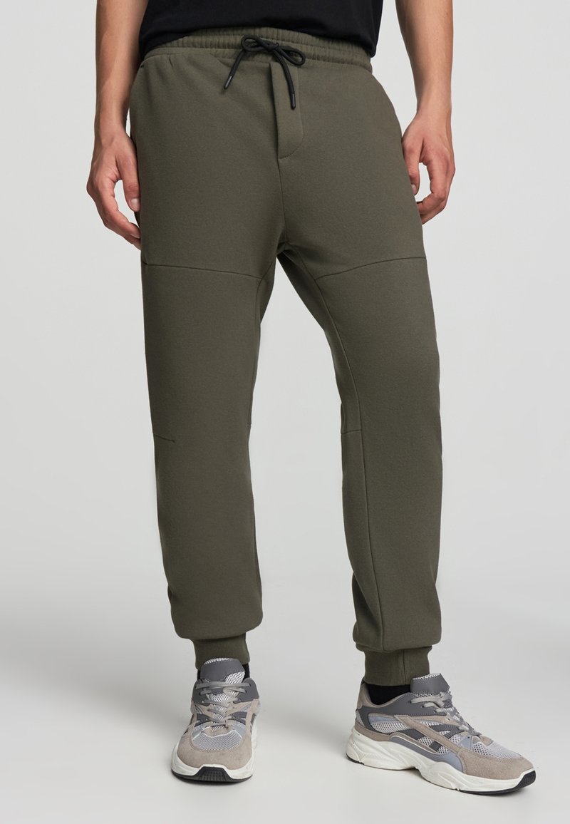 Oliwkowe dresy z elastycznym pasem i regulowanym sznurkiem. Posiadają zwężające się nogawki oraz boczne szwy z teksturowanymi panelami.