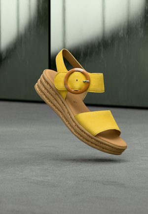 Sandales compensées en cuir jaune avec deux larges brides, une boucle circulaire et une semelle en caoutchouc texturée. Le design met l'accent sur le confort et la stabilité.