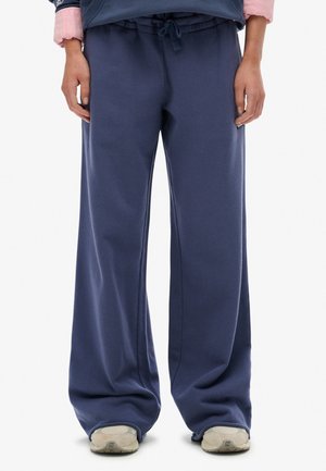 Persona che indossa pantaloni sportivi blu navy larghi e sneaker beige, in piedi davanti a uno sfondo bianco semplice.
