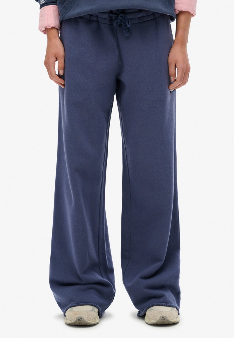 Persona vistiendo pantalones deportivos sueltos de color azul marino y zapatillas beige, de pie contra un fondo blanco liso.