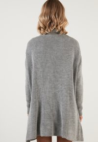 Suéter gris de punto con una textura acanalada, que presenta un corte holgado y un cuello alto. Mangas largas y un detalle de dobladillo asimétrico.