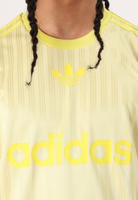 Világos sárga sportpóló függőleges csíkokkal, amelyen egy nagy sárga "adidas" logó és egy háromlevelű embléma található a mellkason.