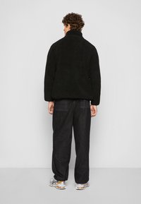 Sort, fluffy fleecejakke med høj krave, oversized pasform og sænkede skuldre. Båret med løse sorte bukser og sølvfarvede sneakers.