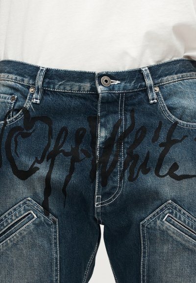 Gros plan sur un jean en denim bleu foncé avec une grande écriture abstraite noire à l'avant et une chemise blanche décontractée rentrée à l'intérieur.