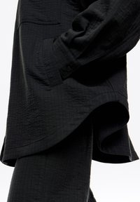 Camisa negra texturizada de corte holgado, con manga abotonada y dobladillo curvado. Incluye un bolsillo en el pecho y un sutil patrón de cuadrícula.