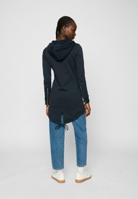 Sweat à capuche bleu marine avec un dos long, des zips sur les côtés et un cordon de serrage ajustable à l'ourlet, associé à un jean bleu clair.