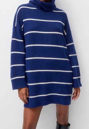 Femme portant une robe-pull à col roulé bleue à manches longues avec des rayures horizontales blanches et des bottines noires, debout sur fond blanc.