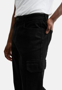Pantaloni cargo in denim nero con vestibilità rilassata, dotati di tasche laterali, chiusura con bottone e dettagli di cucitura a vista. Materiale in cotone.