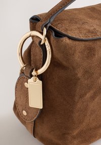 Brown suede handbag with black trim, featuring a large gold ring and attached rectangular gold tag on the side.