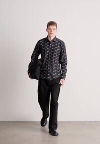 Τυπωμένο μαύρο πουκάμισο με επαναλαμβανόμενο μοτίβο λογότυπου, σε συνδυασμό με μαύρο παντελόνι cargo, και κρατώντας ένα διπλωμένο μπουφάν. Το μοντέλο φοράει μαύρα παπούτσια.