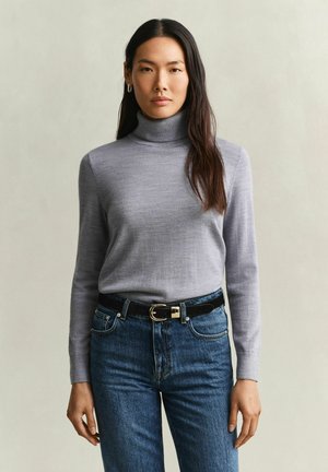 Frau mit langen dunklen Haaren, die einen hellgrauen Rollkragenpullover, blaue Jeans und einen schwarzen Gürtel trägt, steht vor einem einfarbigen Hintergrund.
