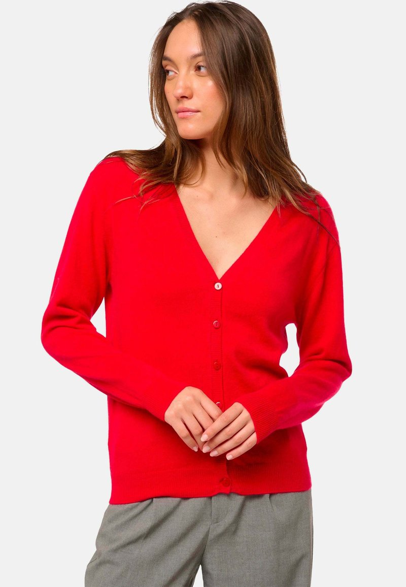 Roter Kardigan mit V-Ausschnitt, langen Ärmeln und Knopfverschluss. Weicher Stoff und taillierte Passform. Kombiniert mit grauen Hosen.