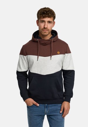 Jonge man met korte krullende haren, draagt een hoodie met kleurvlakken in bruin, wit en zwart met een voorzak en een blauwe spijkerbroek.