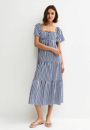 Kobieta ubrana w niebiesko-białą, pionowo prążkowaną sukienkę midi z bufianymi rękawami, stojąca na tle jednolitego białego tła.