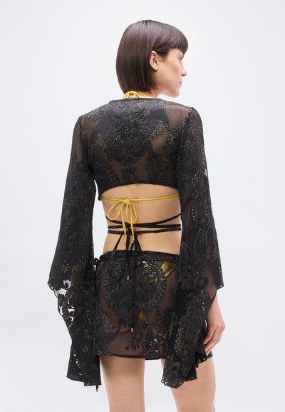 Γυναίκα με κοντά καστανά μαλλιά που φορά διάφανο μαύρο δαντελωτό φόρεμα πάνω από χρυσό μπικίνι, δείχνοντας την πλάτη με κορδόνια και φουσκωτά μανίκια μπροστά σε λευκό φόντο.