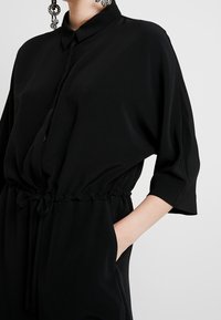 Černý, volně střižený overal s límečkem, krátkými rukávy, elastickým pasem a bočními kapsami. Hladká tkanina s matnou texturou.