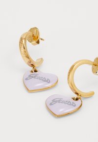 Zlatne kružne naušnice s ljubičastim privjiscima u obliku srca na kojima je sivi "Guess" logotip. Glatka tekstura s poliranim završetkom.