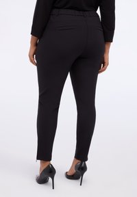 Pantalones ajustados negros con un diseño elegante, que cuentan con una cinturilla elástica y detalles de cremallera en los tobillos, combinados con zapatos de tacón alto negros.