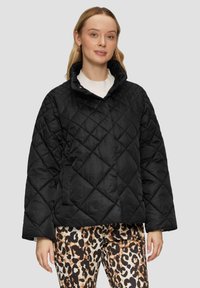 Zwarte, gewatteerde jas met een hoge kraag en een knoopsluiting. Heeft brede mouwen en zijzakken, gecombineerd met een broek met een dierlijk patroon.