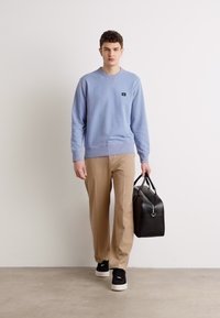 Lyseblå crewneck sweatshirt, beige bukser, sorte sneakers, holder en sort lædertaske, står foran en ensfarvet baggrund.